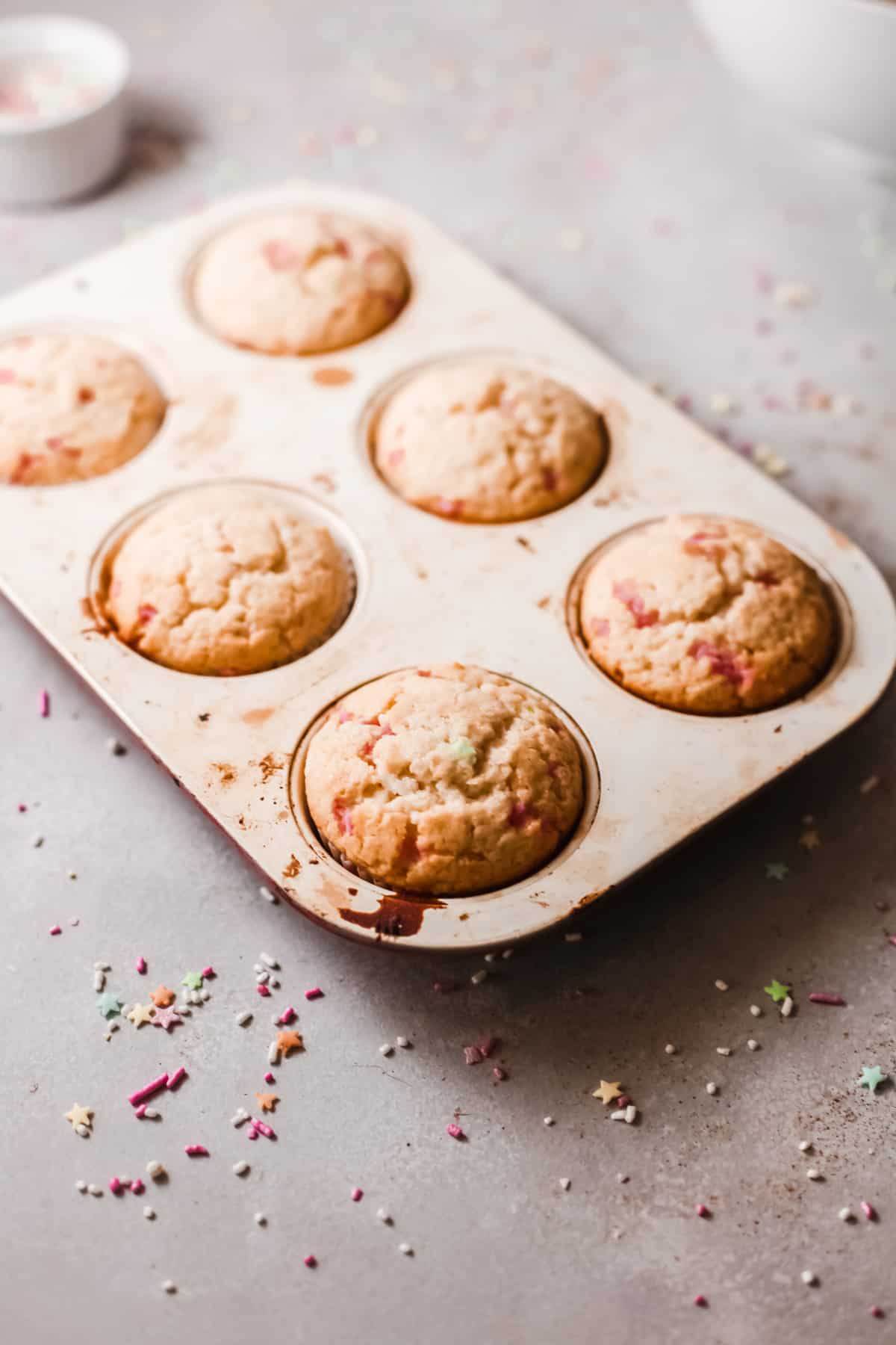 Six baked party cupcakes in a muffin tin waiting to be frosted.