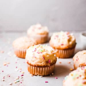 Six birthday cupcakes topped with vanilla buttercream frosting and sprinkles on a gray countertop.