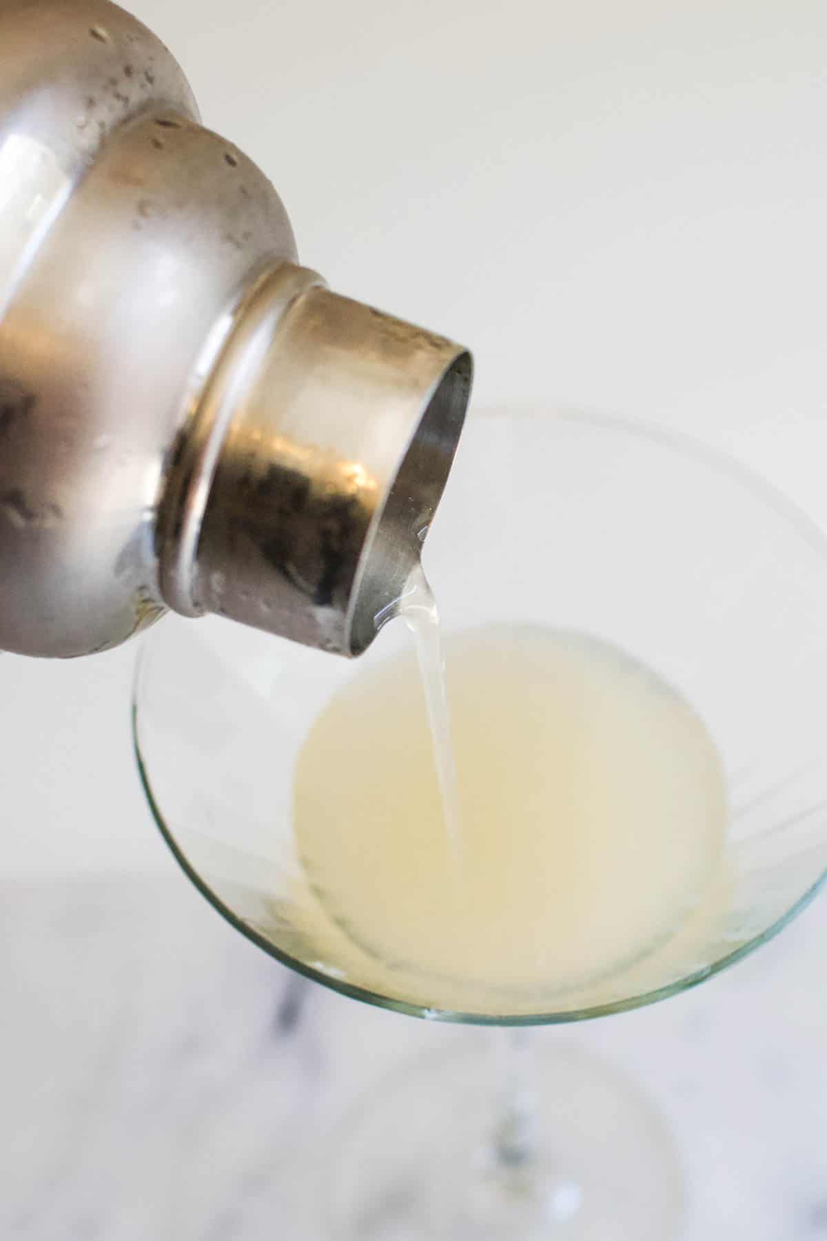 A cocktail shaker straining the French gimlet into a martini glass.