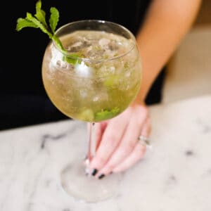 A woman with her hand on the stem of a spritz glass on a counter holding a Hugo Spritz.