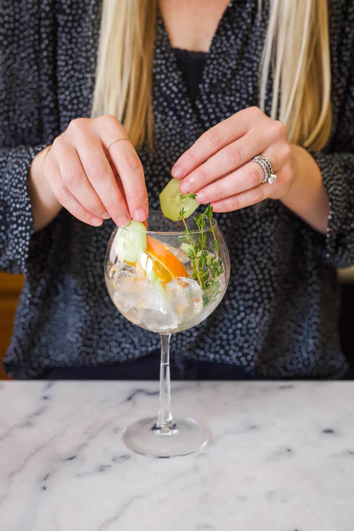 A woman adding cucumber slices to a wine glass filled with ice and citrus slices.