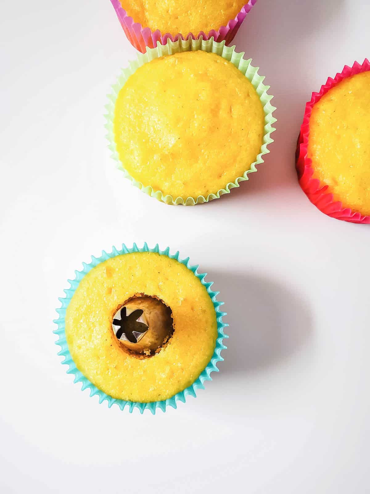 Baked cupcakes on a table, one has a the wide end of a piping tip stuck in the center to poke an easy hole.