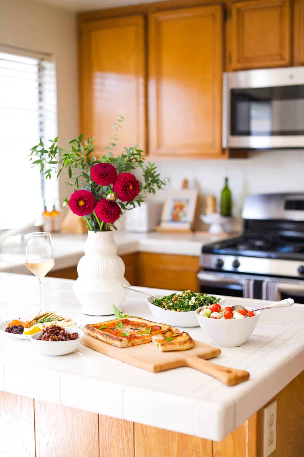 White serving dishes holding food on a kitchen counter with a vase of flowers and a wine glass.