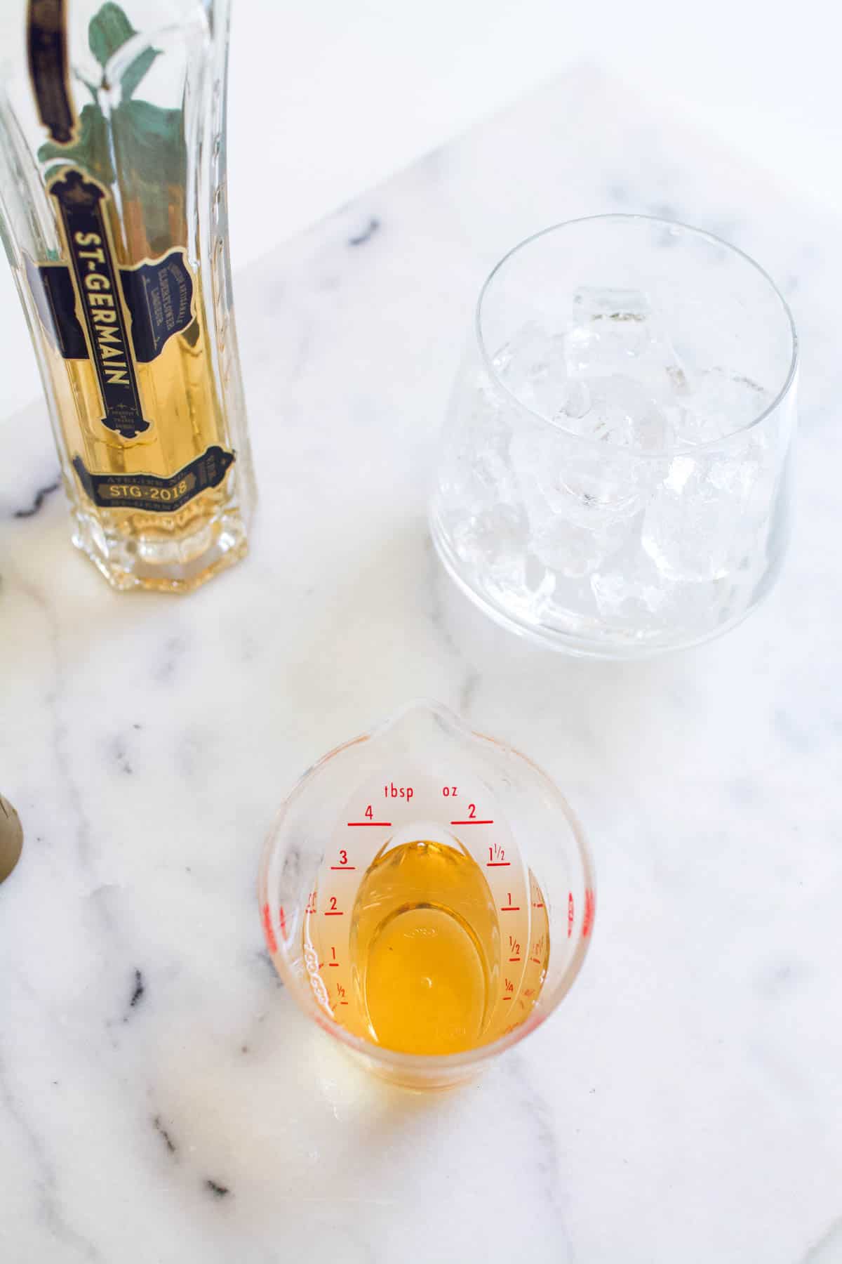 A small plastic measuring cup filled with St. Germain liqueur in front of a glass filled with ice and a bottle of St. Germain.