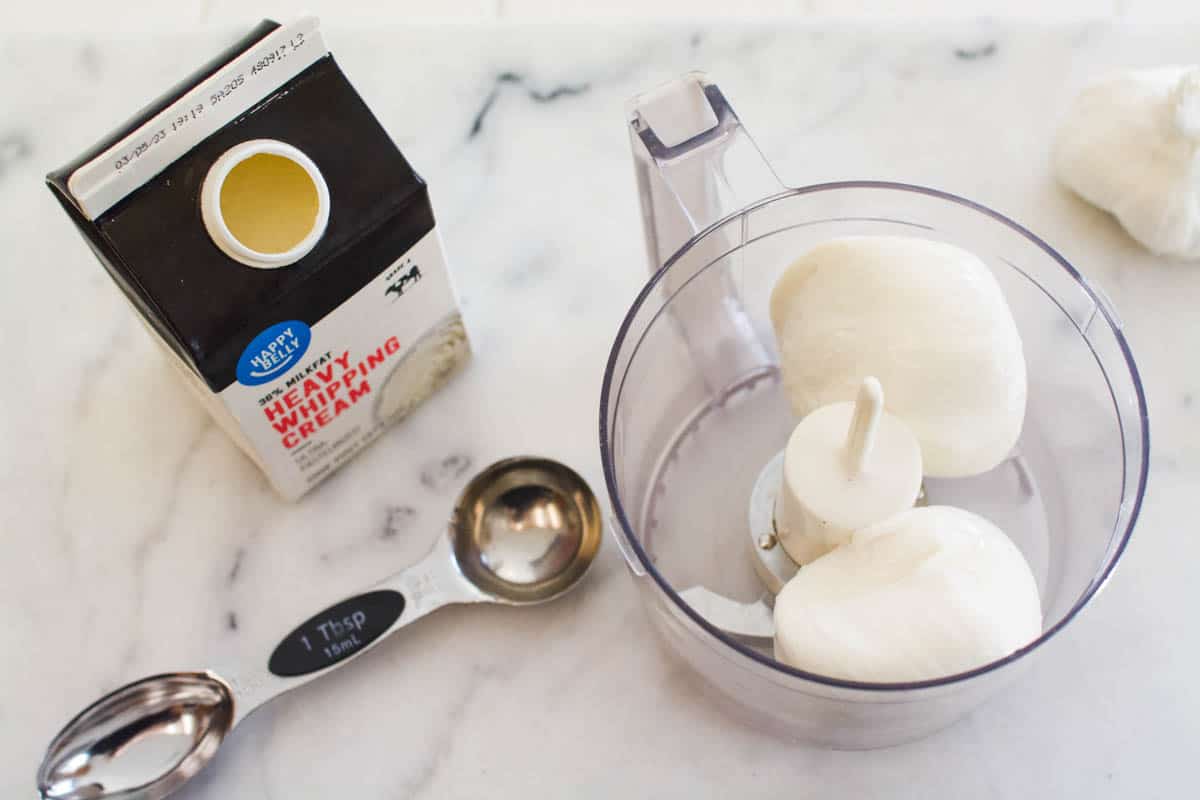 A food processor bowl filled with burrata, next to a measuring spoon and small carton of heavy whipping cream.