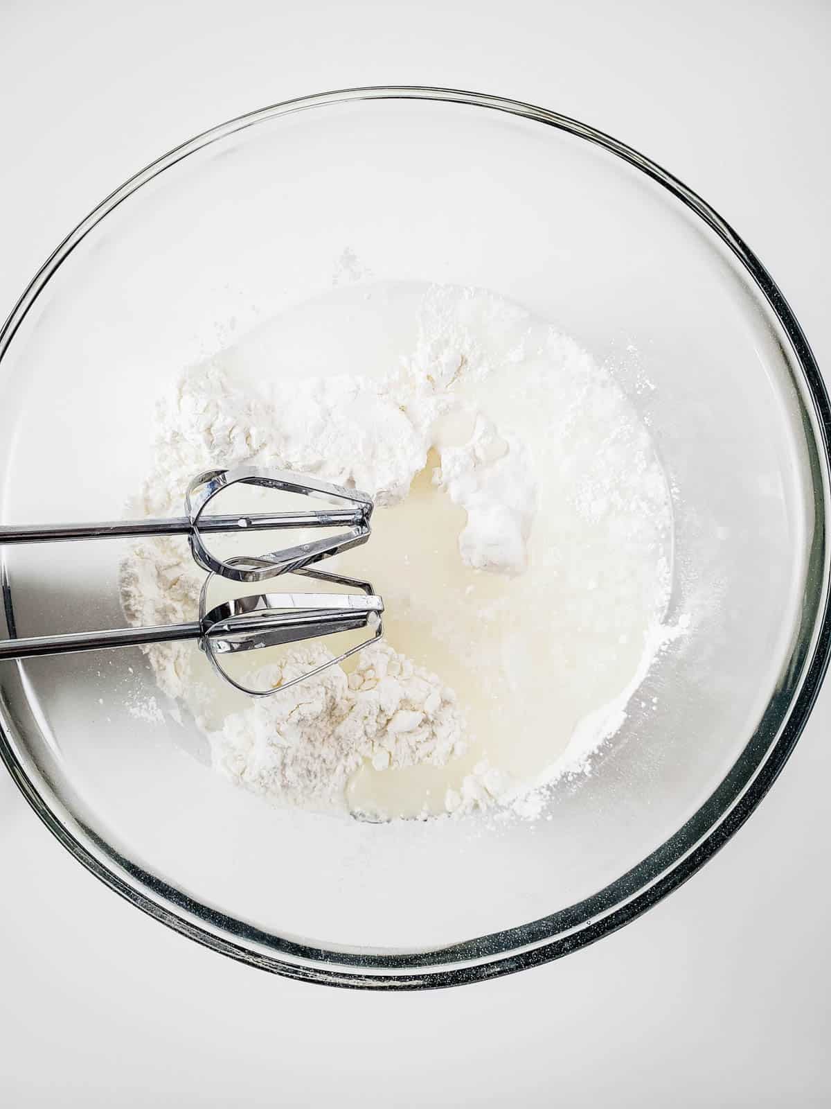 A glass bowl with angel food cake mix and water and a handheld mixer.