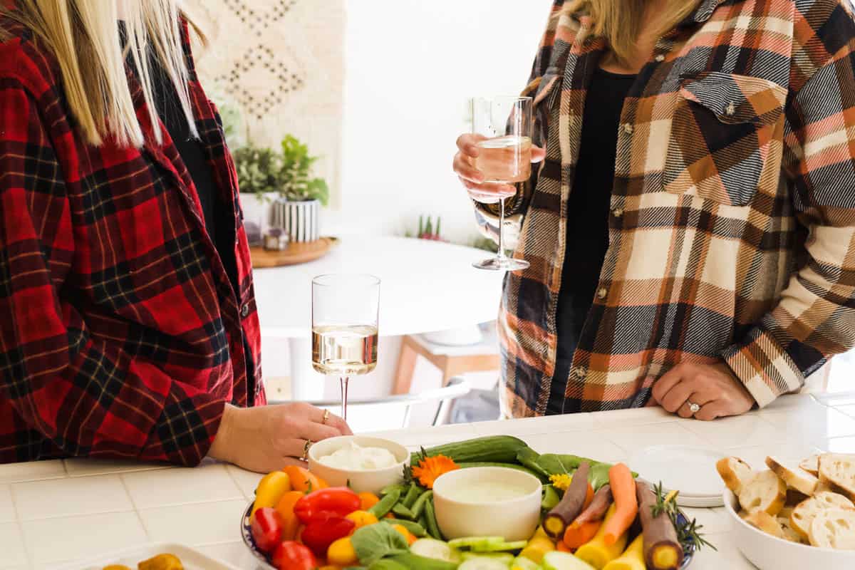 Two friend hanging out with glasses of wine and dips and veggies next to them.