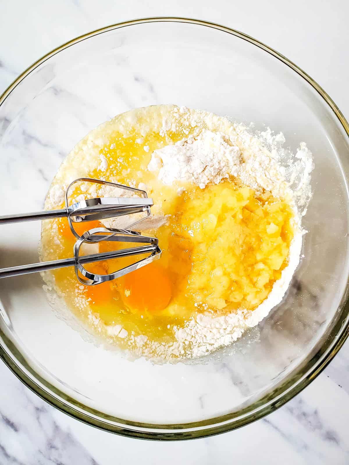 Large glass mixing bowl with the ingredients and a hand mixer to make pineapple cupcakes.