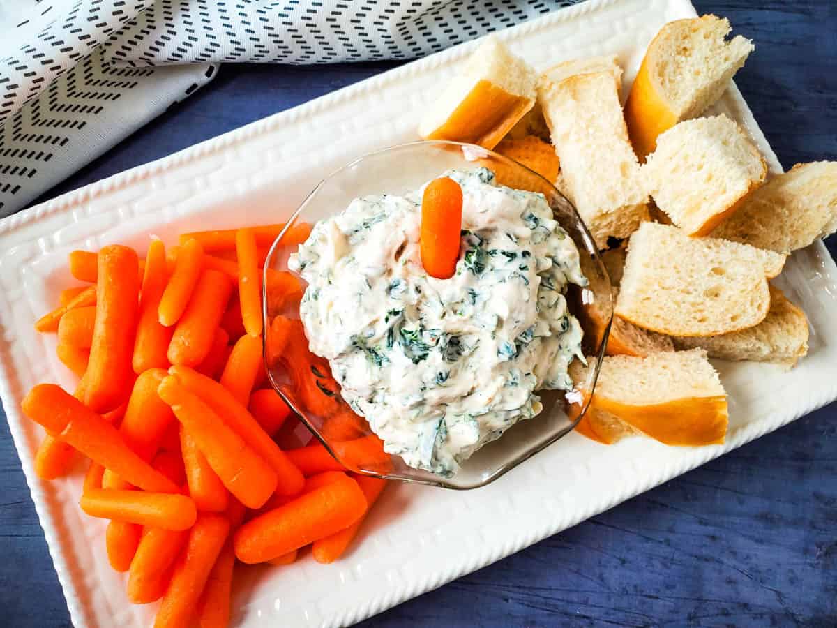 Cold spinach dip in a glass bowl atop a white serving dish with carrots and bread.