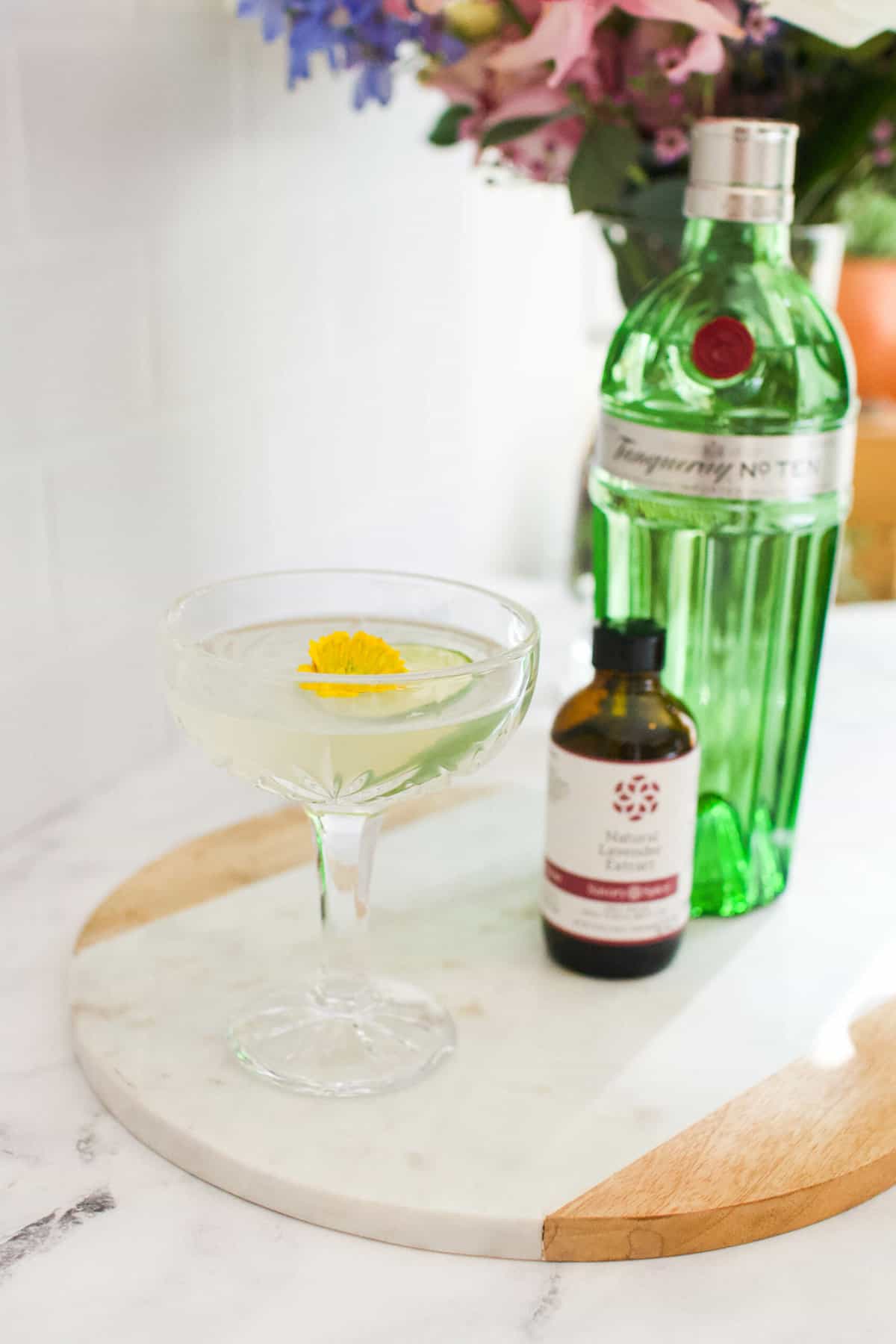 Lavender gimlet on a wood and marble board with bottles of gin and lavender bitters. There is a vase of flowers in the background.