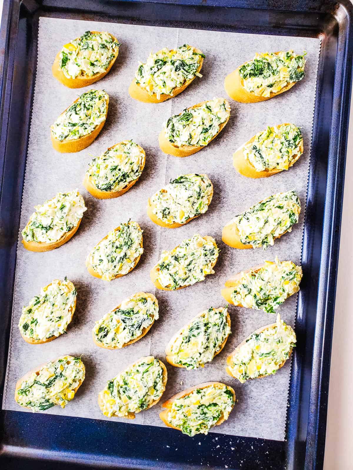 A cookie sheet with sliced baguette spread with spinach artichoke dip before baking.