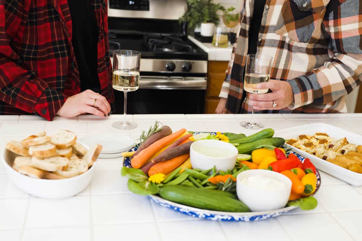 A try with veggies and dips for a girl's night in with woman and wine glasses behind it.