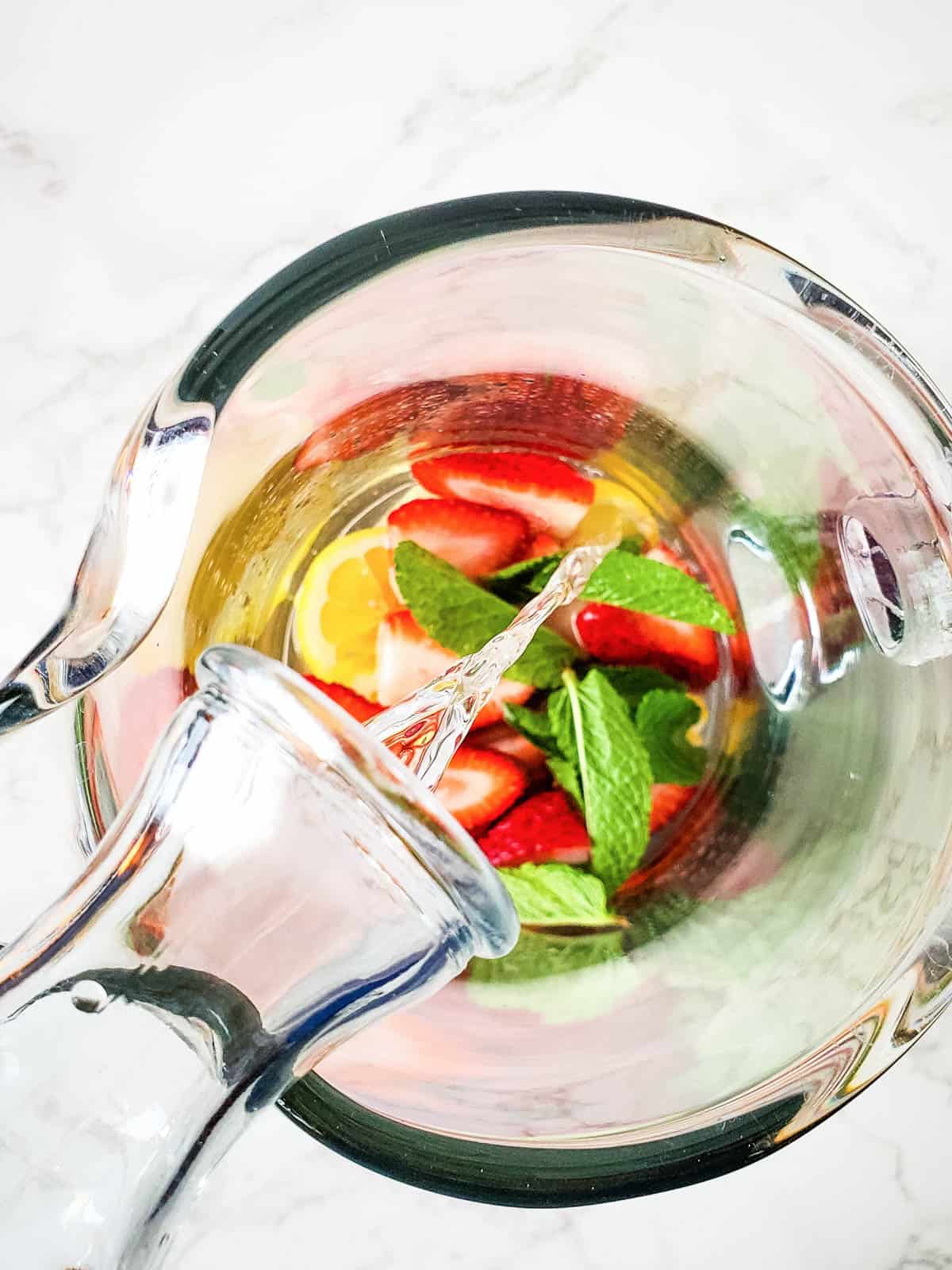 Pouring vodka into a glass pitcher of cut fruit. 