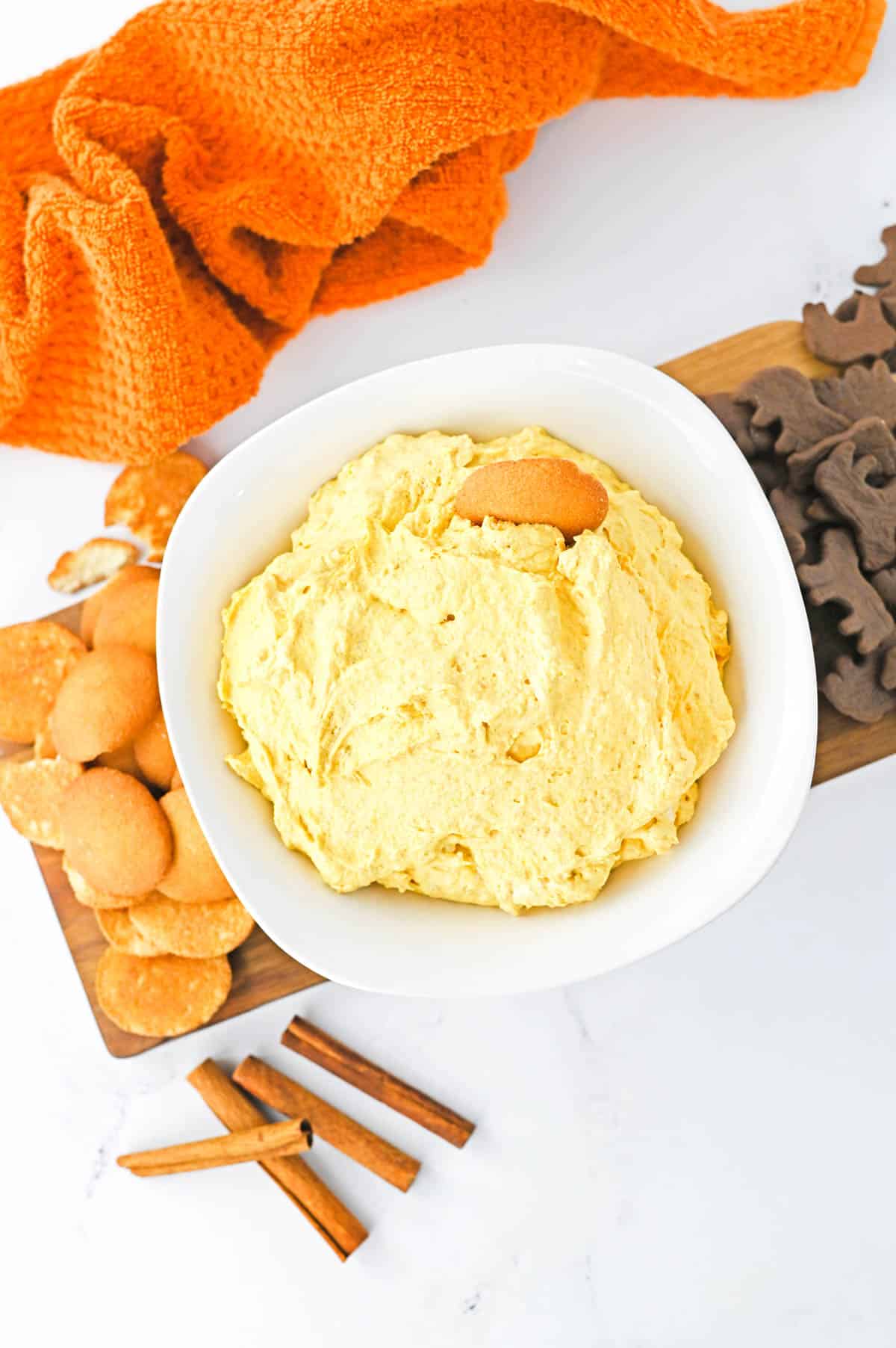 An orange towel on a table next to a wood tray with cookies and white bowl with pumpkin dip.