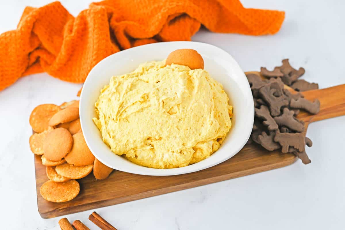 A wooden tray on a table topped with cookies and a bowl holding pumpkin dessert dip.