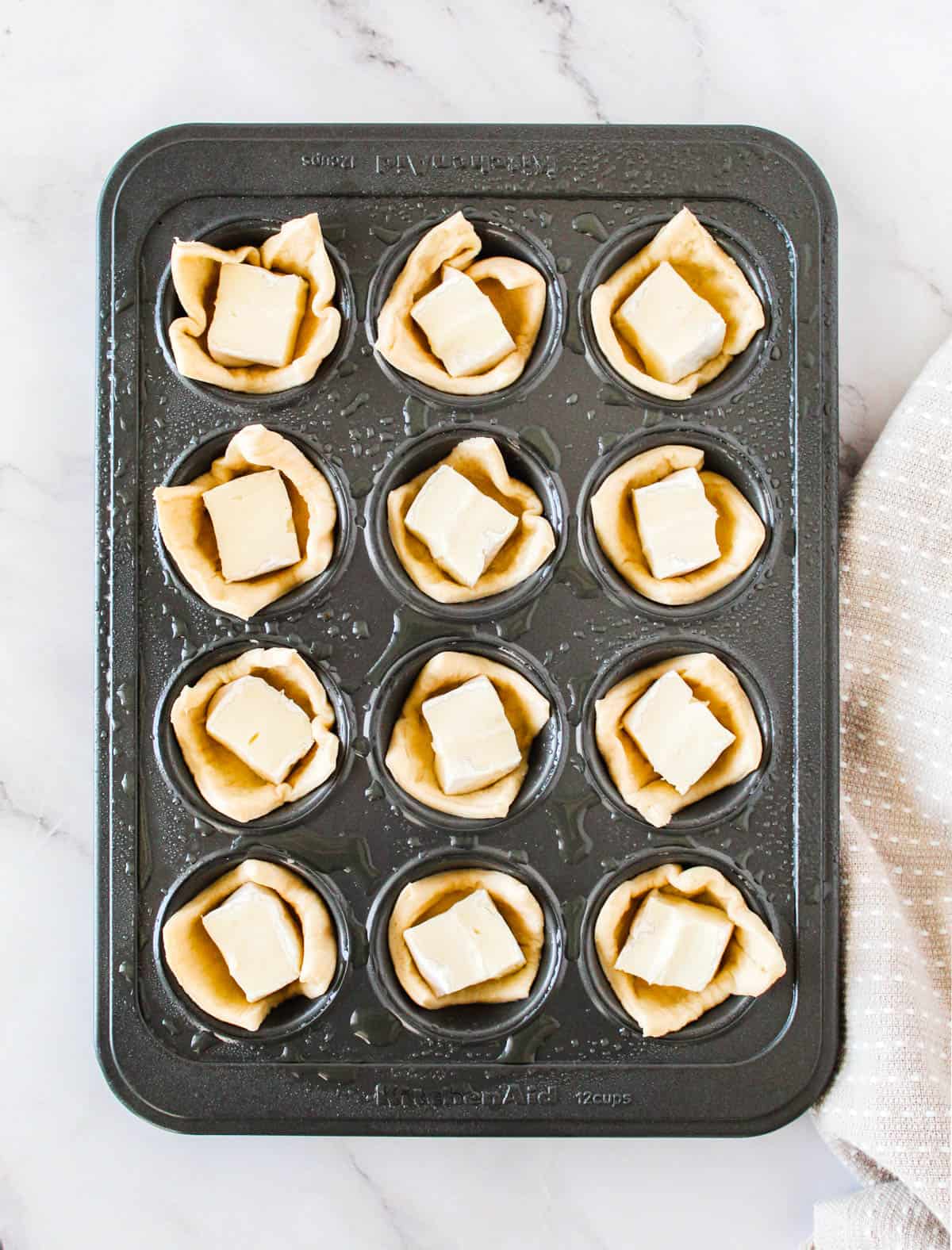 Crescent rolls pressed on mini muffin tin slots each filled with a block of brie cheese.