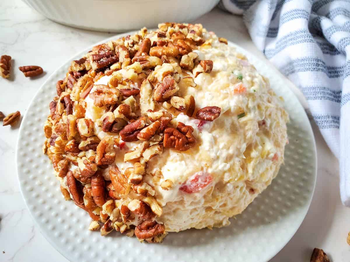 Cheese ball with pineapple, peppers, green onion, and pecans on a white plate.