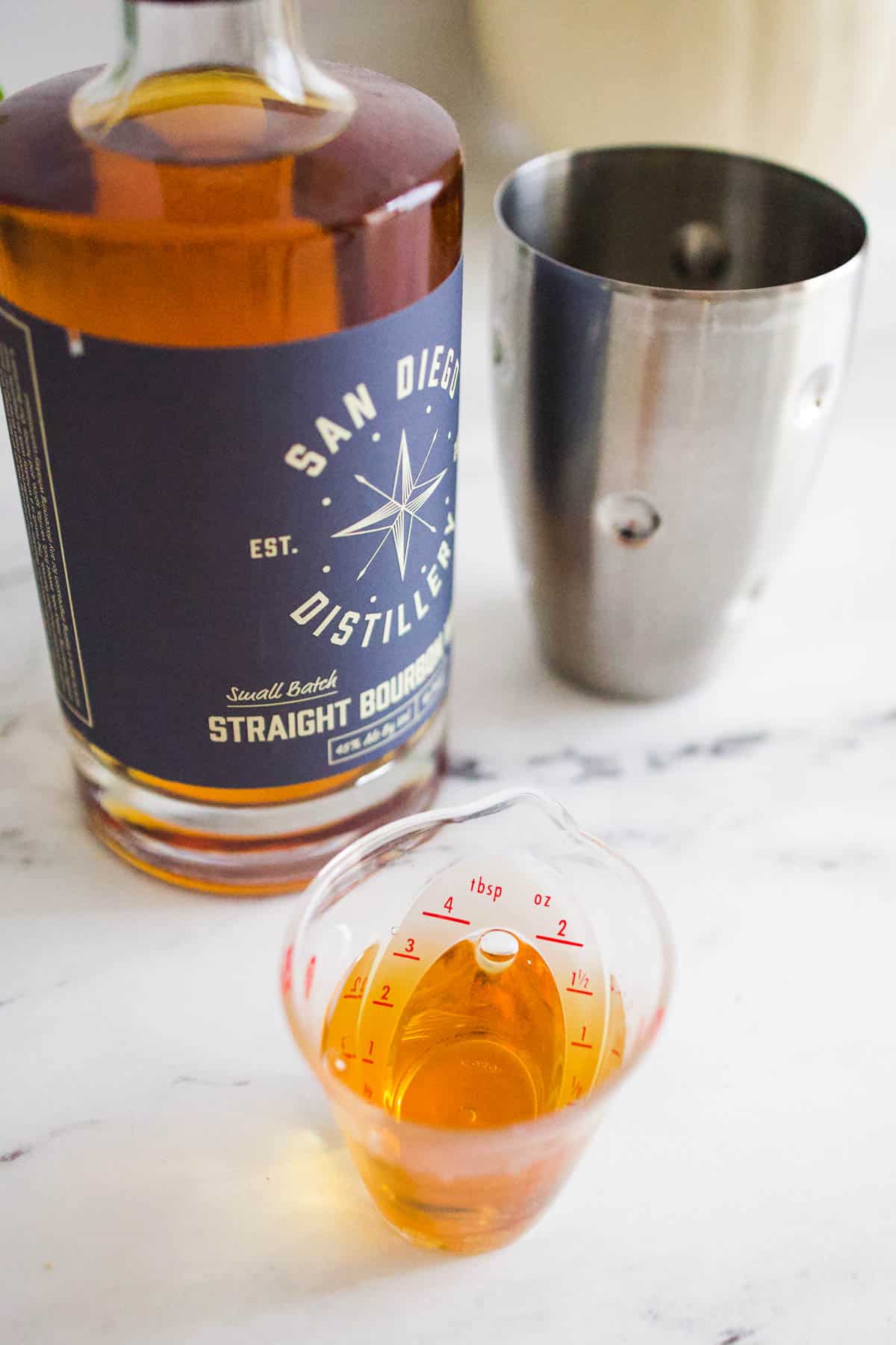 A small measuring cup with bourbon next to a bottle of bourbon and a cocktail shaker.