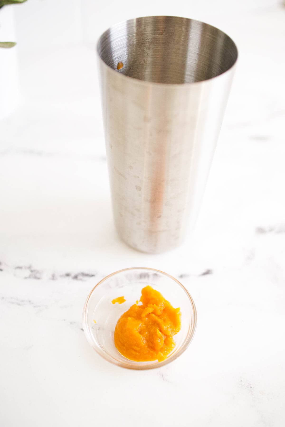 A small bowl with pumpkin puree on a table next to a cocktail shaker.