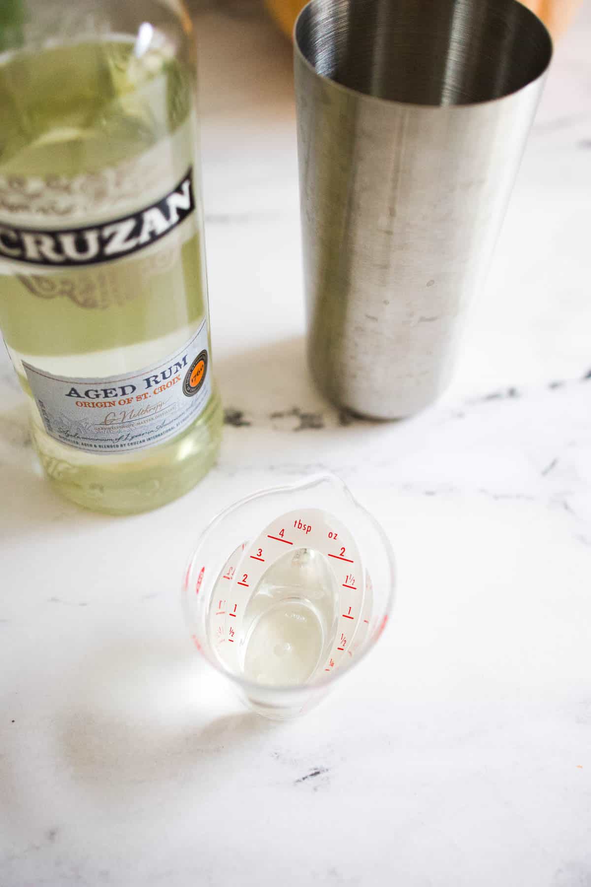 Rum in a measuring cup next to a bottle of rum and a cocktail shaker.