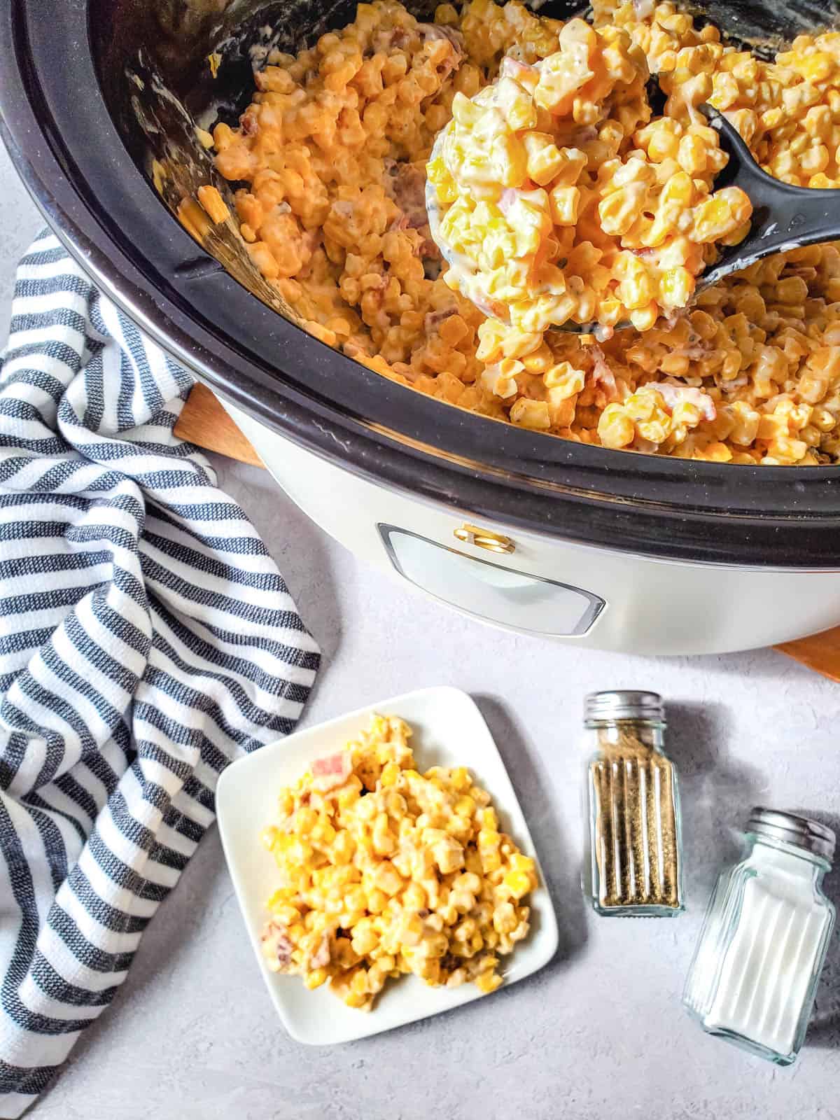 A slow cooker with cream cheese corn and bacon on a table with a portion of the recipe on a small white plate next to it.