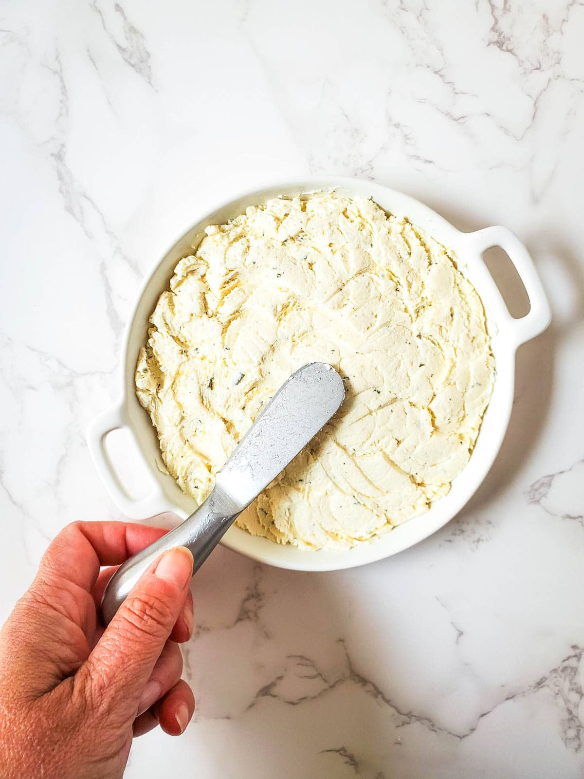 A woman using a cheese spreader to spread out the top of boursin cheese in a serving dish.