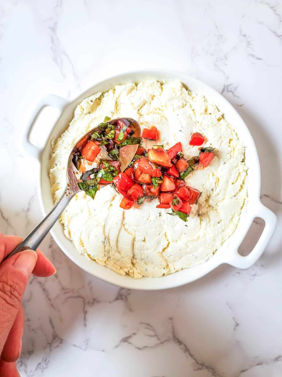 A white serving dish holding boursin being topped with a bruschetta topping by a woman holding a spoon.