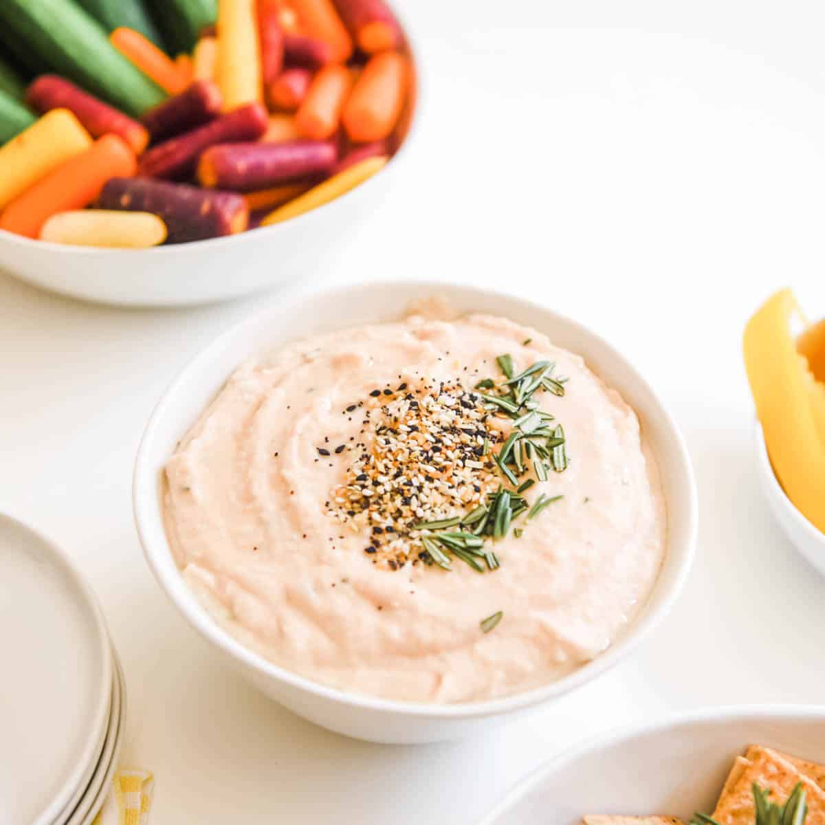 White bean dip in a white bowl on a table next to vegetables.