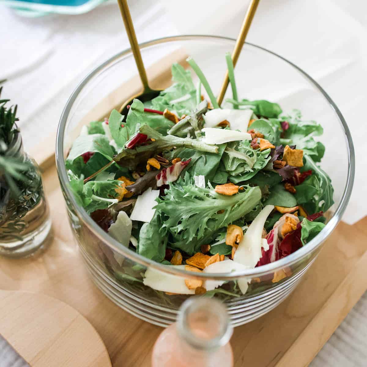 A green salad in a bowl with cheese and serving spoons.