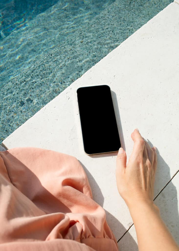 Woman reaching for a phone while sitting next to a swimming pool.